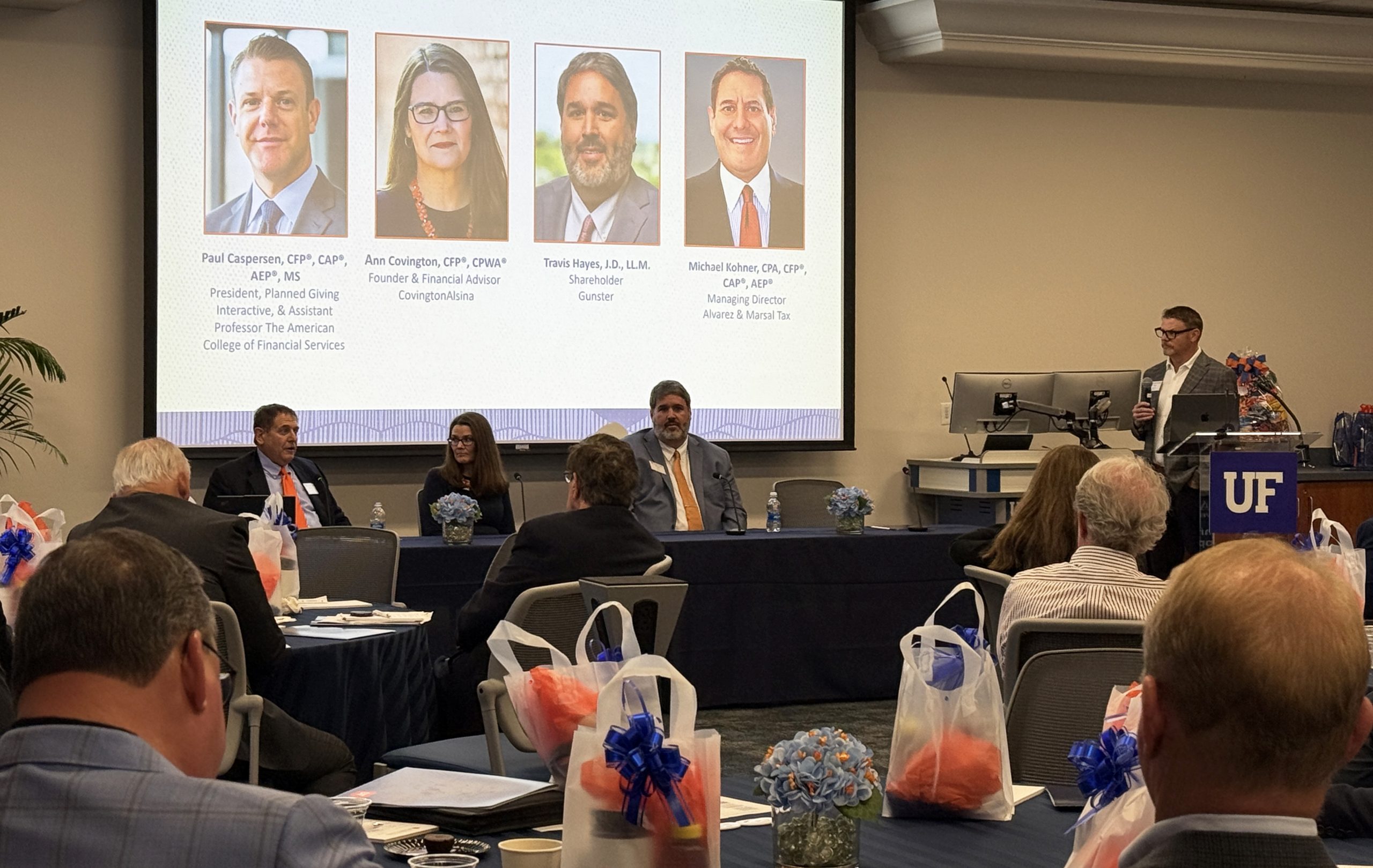 The image is a photo of a panel of advisors presenting at the UFAN Gator Gathering event. The panelists, Michael Kohner, Ann Covington and Travis Hayes sit behind a long table and Paul Caspersen is at the podium with a microphone. There is an audience listening to them present.